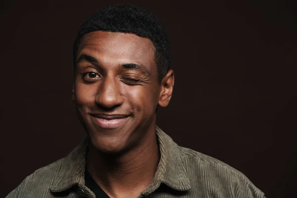 Young black man dressed in shirt smiling and winking at camera isolated over brown wall