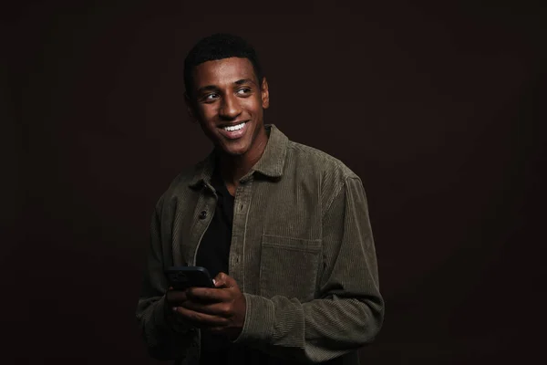 茶色の壁の上に隔離された携帯電話を使用している間 若い黒人男性は笑っている — ストック写真