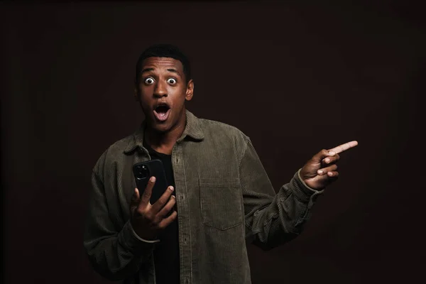 Shocked black man pointing finger aside while using cellphone isolated over brown wall