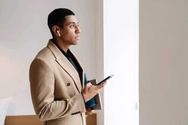 Black man wearing suit using cellphone and wireless earphones indoors