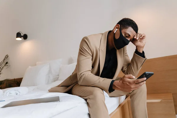 Black man in face mask using cellphone and wireless earphones while sitting on bad in hotel