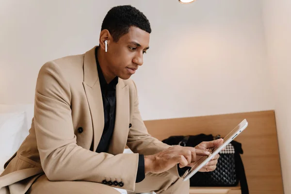 Black man using tablet computer and wireless earphones while sitting on bad in hotel