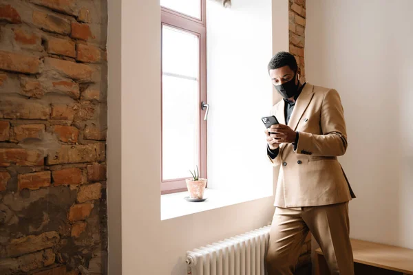 Black man in face mask using wireless earphones and cellphone by window indoors