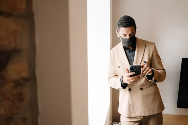 Black man in face mask using wireless earphones and cellphone by window indoors