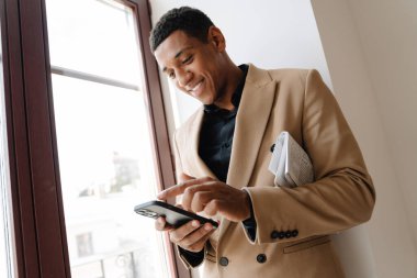 Takım elbiseli siyah bir adam pencerenin önünde beklerken cep telefonu kullanıyor.