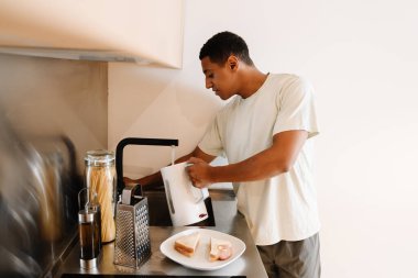 Otelde kahvaltı hazırlarken elinde çaydanlık tutan tişörtlü siyah bir adam.