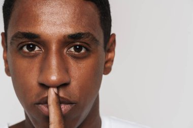 Genç siyah adam beyaz duvarın üzerinden izole edilmiş kameraya sessiz jest yapıyor.