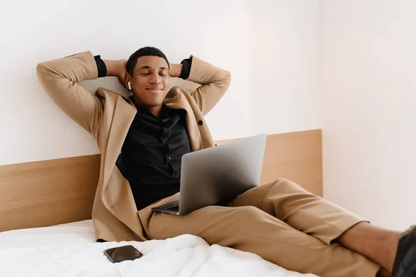 Black man in earphones smiling and using laptop while sitting on bad at hotel