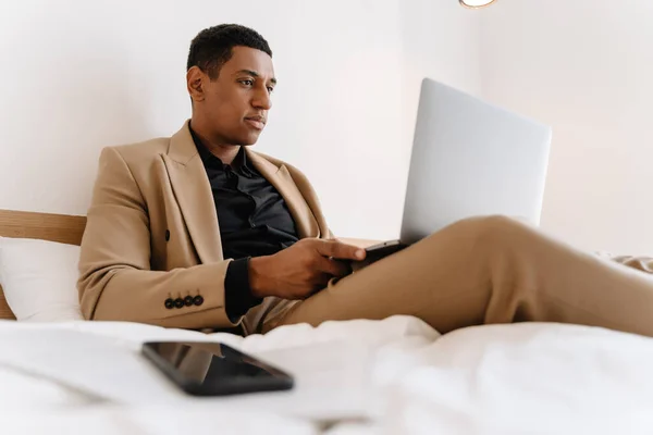 Black man working on laptop while sitting on bad at hotel