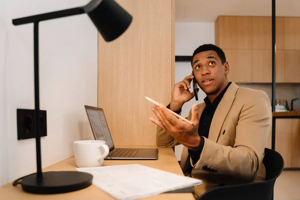 Black man talking on cellphone while working with laptop at desk in hotel