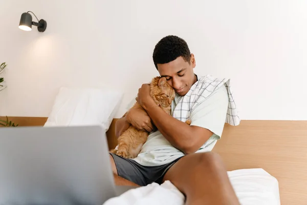 Black man wearing t-shirt petting his cat while working with laptop in hotel