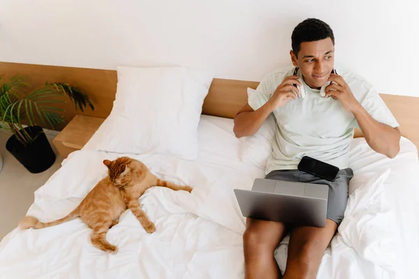 Black man smiling and using laptop while sitting with his cat on bad in hotel