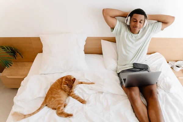 Black man in headphones using laptop while sitting with his cat on bad in hotel
