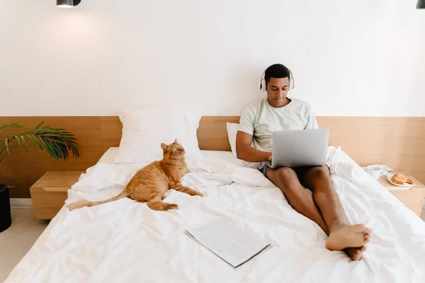 Black man in headphones using laptop while sitting with his cat on bad in hotel