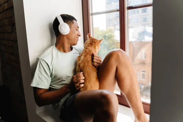 Black man in headphones sitting with his cat on windowsill in hotel