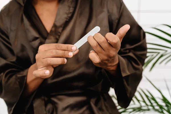 Black man wearing bathrobe doing manicure with nail file in bathroom