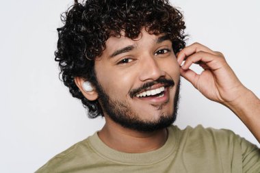 Young middle eastern man laughing while listening music with earphones isolated over white wall