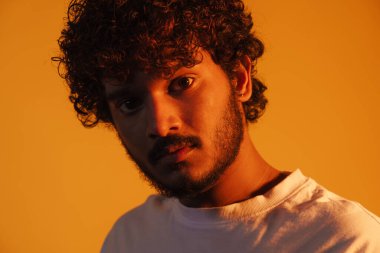 Young bearded man dressed in t-shirt posing and looking at camera isolated over yellow background