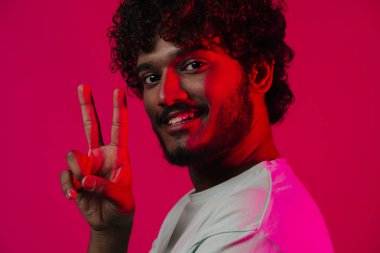 Orta Doğulu genç adam gülümsüyor ve pembe duvarda izole edilmiş barış işareti gösteriyor.