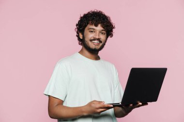 Orta Doğulu genç adam pembe duvarın üzerinde dizüstü bilgisayarla poz verirken gülümsüyor.