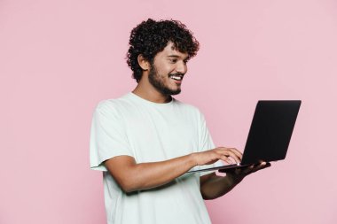 Orta Doğulu genç adam pembe duvarın üzerinde dizüstü bilgisayarla poz verirken gülümsüyor.