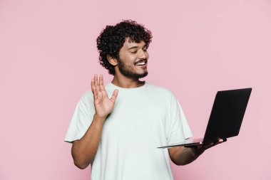 Orta Doğulu genç adam pembe duvarın üzerinde dizüstü bilgisayarla poz verirken el sallıyor.
