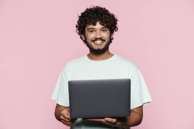 Young middle eastern man smiling while posing with laptop isolated over pink wall