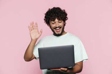Orta Doğulu genç adam pembe duvarın üzerinde dizüstü bilgisayarla poz verirken el sallıyor.