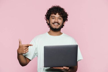Orta Doğulu genç adam gülümserken pembe duvarın üzerinde dizüstü bilgisayarını işaret ediyor.