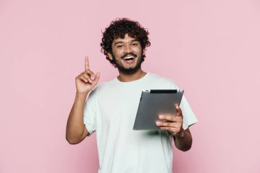 Tablet bilgisayarlı genç, orta doğulu bir adam pembe duvarın üzerinde parmağını yukarı doğru gösteriyor.
