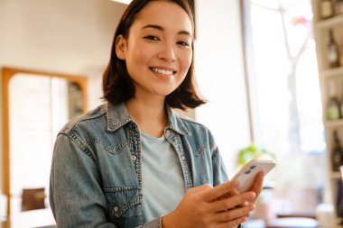 Kafede gülümseyen ve cep telefonu kullanan Asyalı genç bir kadın.
