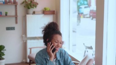 Afro saçlı neşeli bir kadın kafede otururken telefonla konuşuyor.