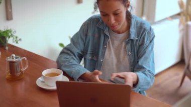 Afro saçlı, gülümseyen bir kadın telefonda yazı yazıyor ve kafede otururken dizüstü bilgisayara bakıyor.