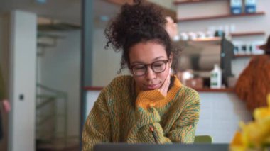 Pleased woman wearing yellow sweater and eyeglasses working on laptop and looking around in cafe