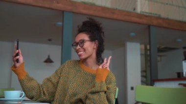 Happy woman wearing yellow sweater and eyeglasses making selfie by phone in cafe