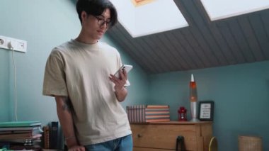 Neşeli Asyalı adam akşamları evde yatağın yanında dikilirken telefonla yazı yazıyor.