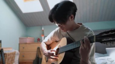 Pretty Asian man plays the guitar while sitting on the bed at home