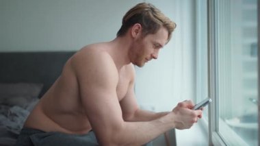 Çıplak gövdesiyle gülümseyen sarışın atletik adam telefonla mesaj atıyor ve eve bakıyor.