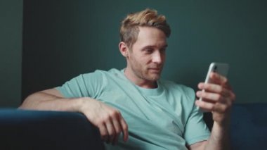 Mutlu sarışın atletik adam cep telefonuyla yazı yazıyor ve evde otururken yan tarafa bakıyor.