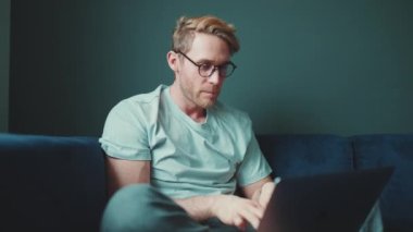Gözlüklü, evde dizüstü bilgisayarla çalışan konsantre sarışın atletik adam.
