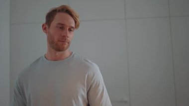 Serious blond athletic man walking and drinking orange juice in the kitchen at home