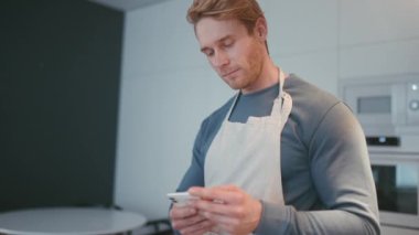 Positive athletic man wearing apron typing on mobile in the kitchen at home