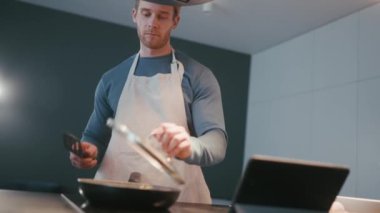 Handsome athletic man mixing frozen vegetables on a frying pan and typing on phone in the kitchen at home