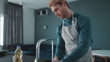 Serious athletic blond man wearing apron washes dishes in the kitchen at home