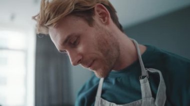 Positive athletic blond man wearing apron washes dishes in the kitchen