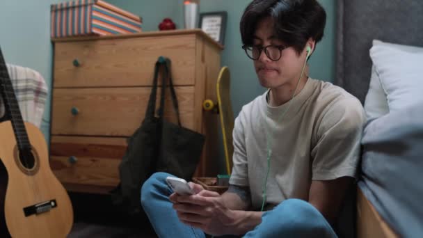 Smiling Asian Man Typing Smartphone Headphones While Sitting Floor Bed — Stockvideo