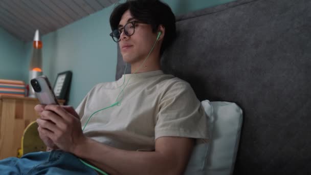 Χαλαρωμένος Ασιάτης Πληκτρολογώντας Στο Smartphone Ακουστικά Ενώ Βρίσκεται Στο Κρεβάτι — Αρχείο Βίντεο