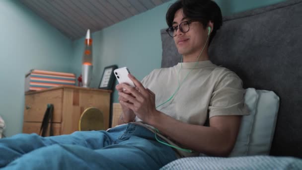 Positive Asian Man Typing Smartphone Headphones While Lying Bed Home — Vídeo de Stock