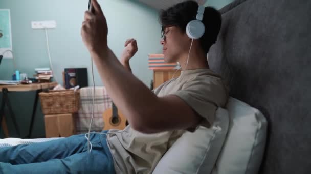 一个英俊的亚洲男人躺在床上用耳机听音乐 — 图库视频影像