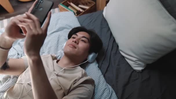 Smiling Asian Man Listening Music Headphones Looking Phone While Lying — Stok Video